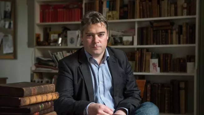 Photo of Will Poole sitting at a desk. There is a pile of early modern books on the desk in front of him, and bookshelves behind him. 